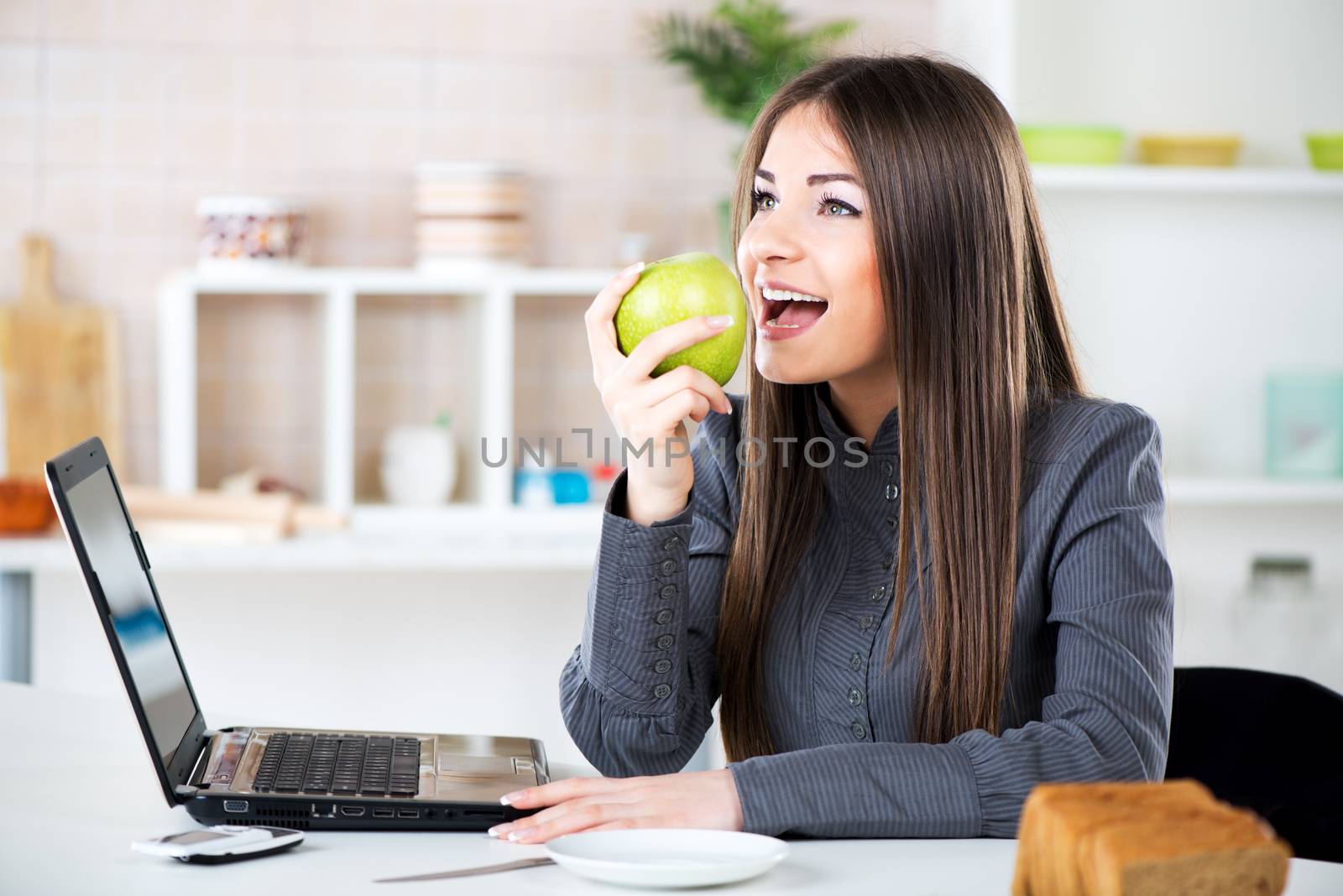 Businesswoman in the kitchen eating apple and reading mail on laptop before going to work.