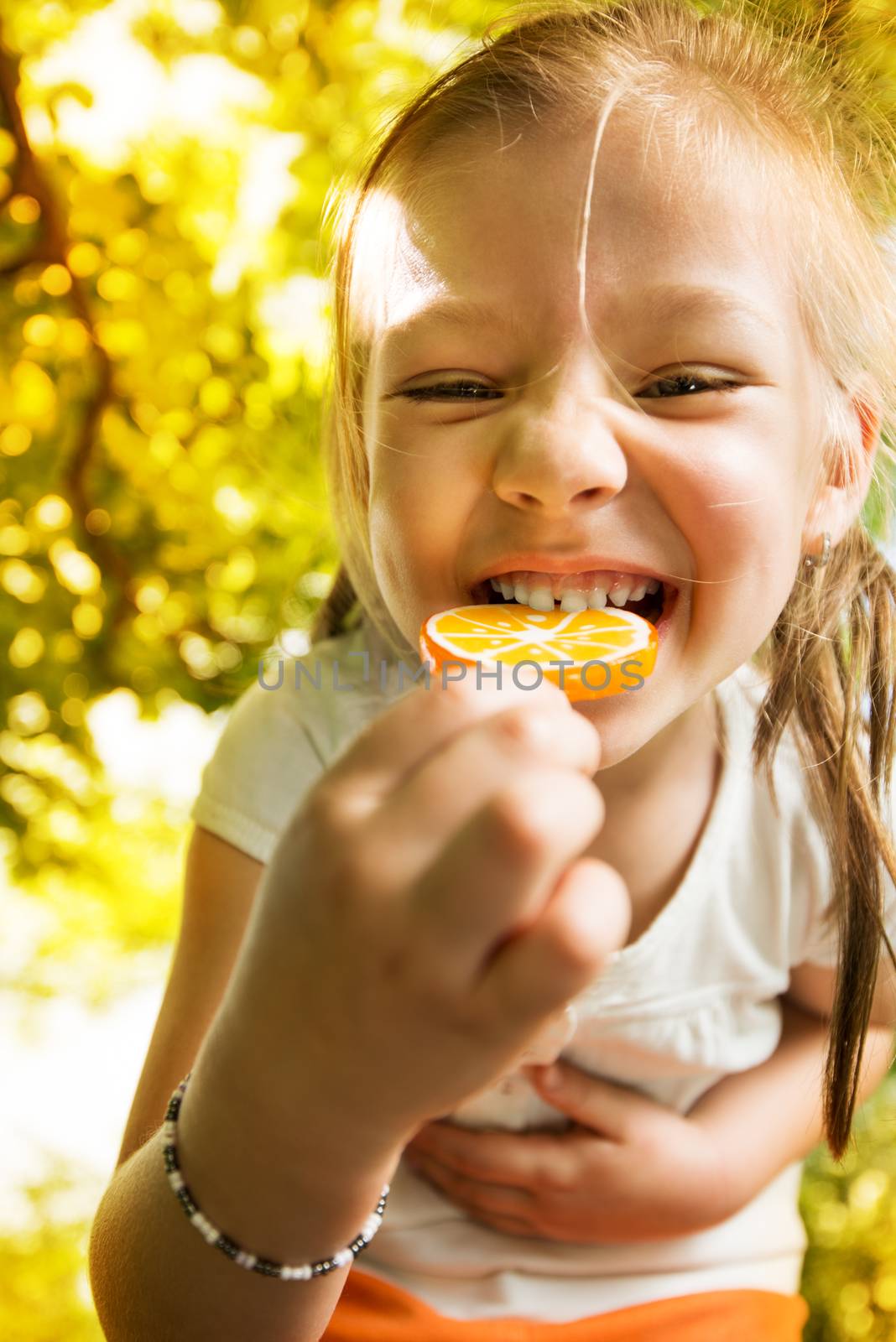 Portrait of Cute Little Girl Standing in the Park and holding in hand orange Lollipop.