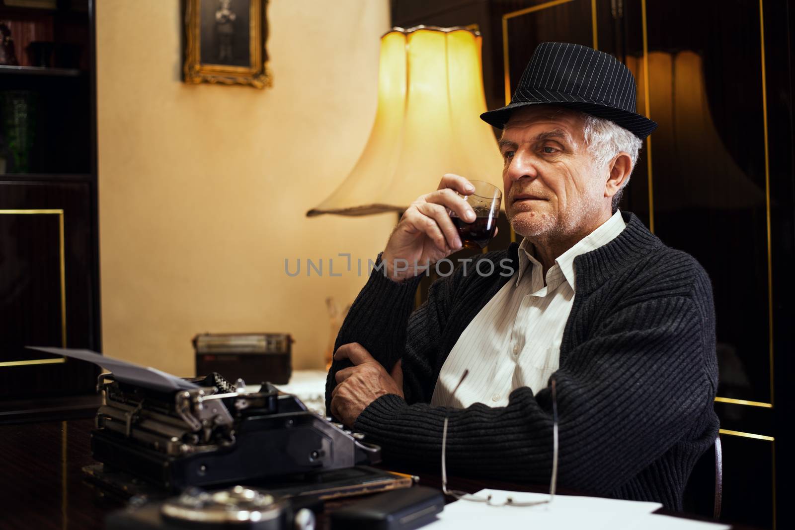 Retro Senior man writer with hat, sitting at the desk with glass of whiskey and thinking.