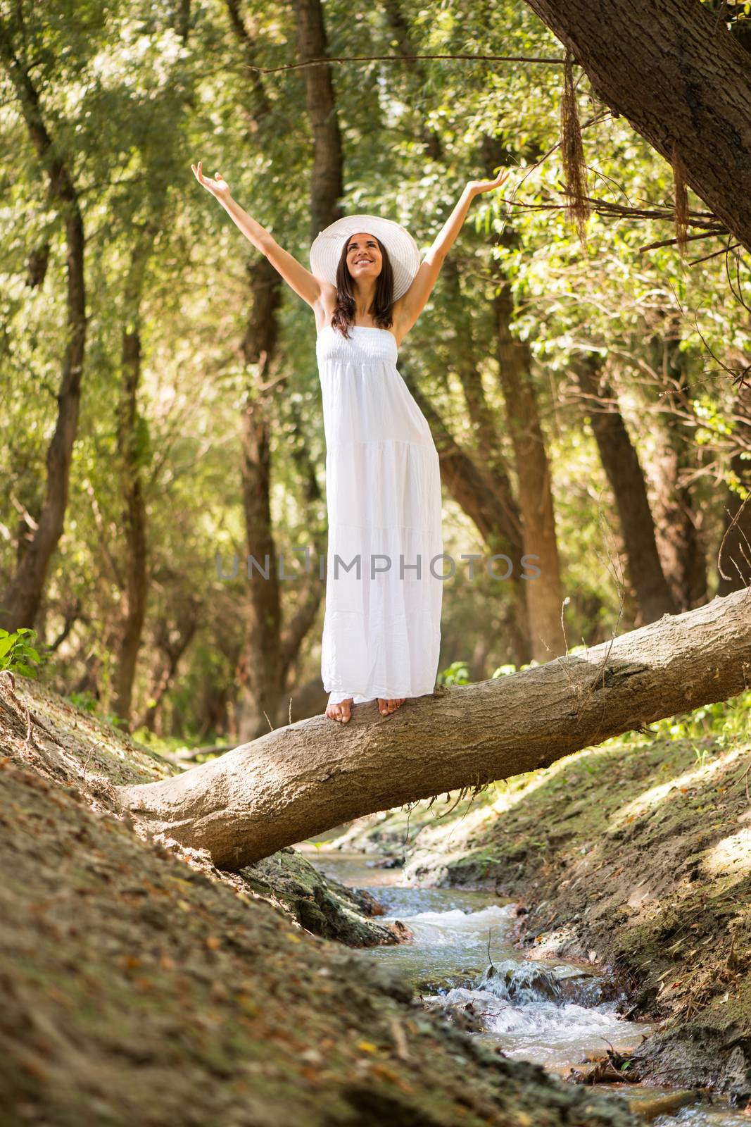Young beautiful woman with arms raised in white dress standing on a tree across the creek.