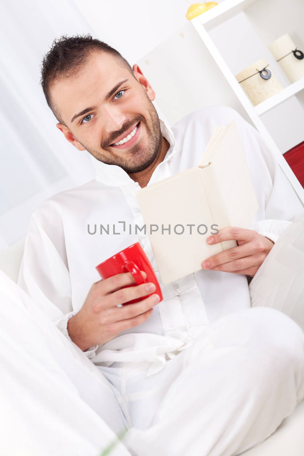 Beautiful young man resting with book and drinking coffee at home.