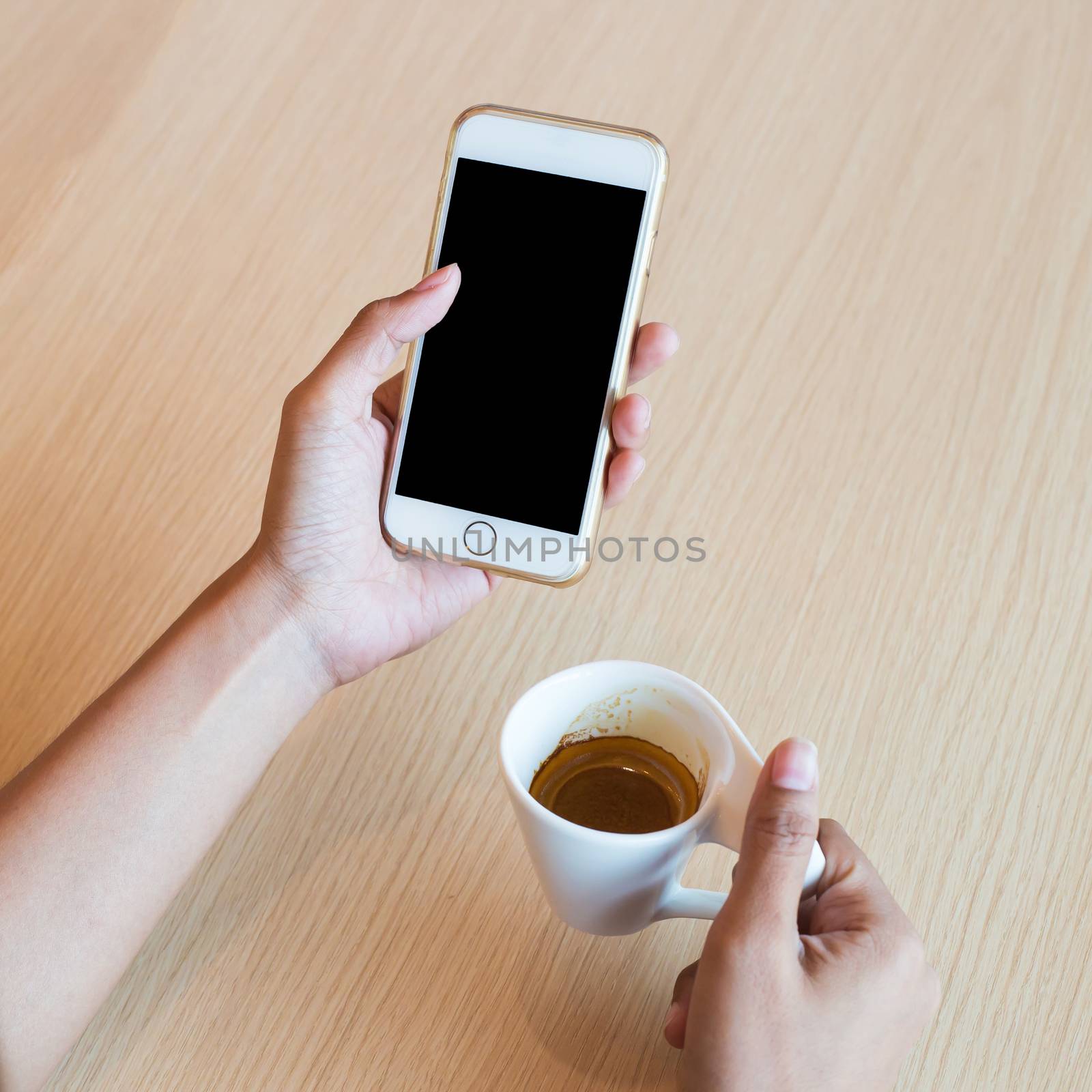Hand using a touch screen of white smartphone on the wooden table and hand holding aromatic cappuccino, white coffee cup, close up