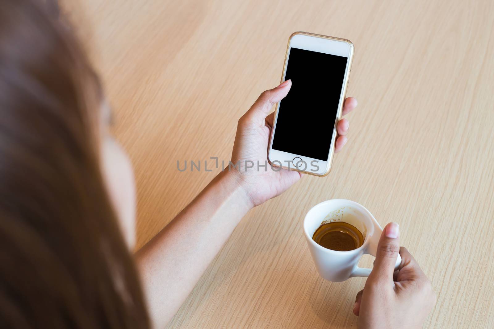 Woman using a touch screen of white smartphone on the wooden table and hand holding aromatic cappuccino, white coffee cup, close up