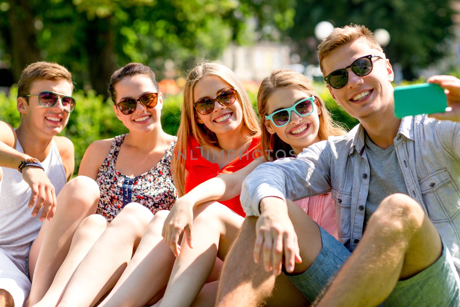 smiling friends with smartphone sitting on grass by dolgachov