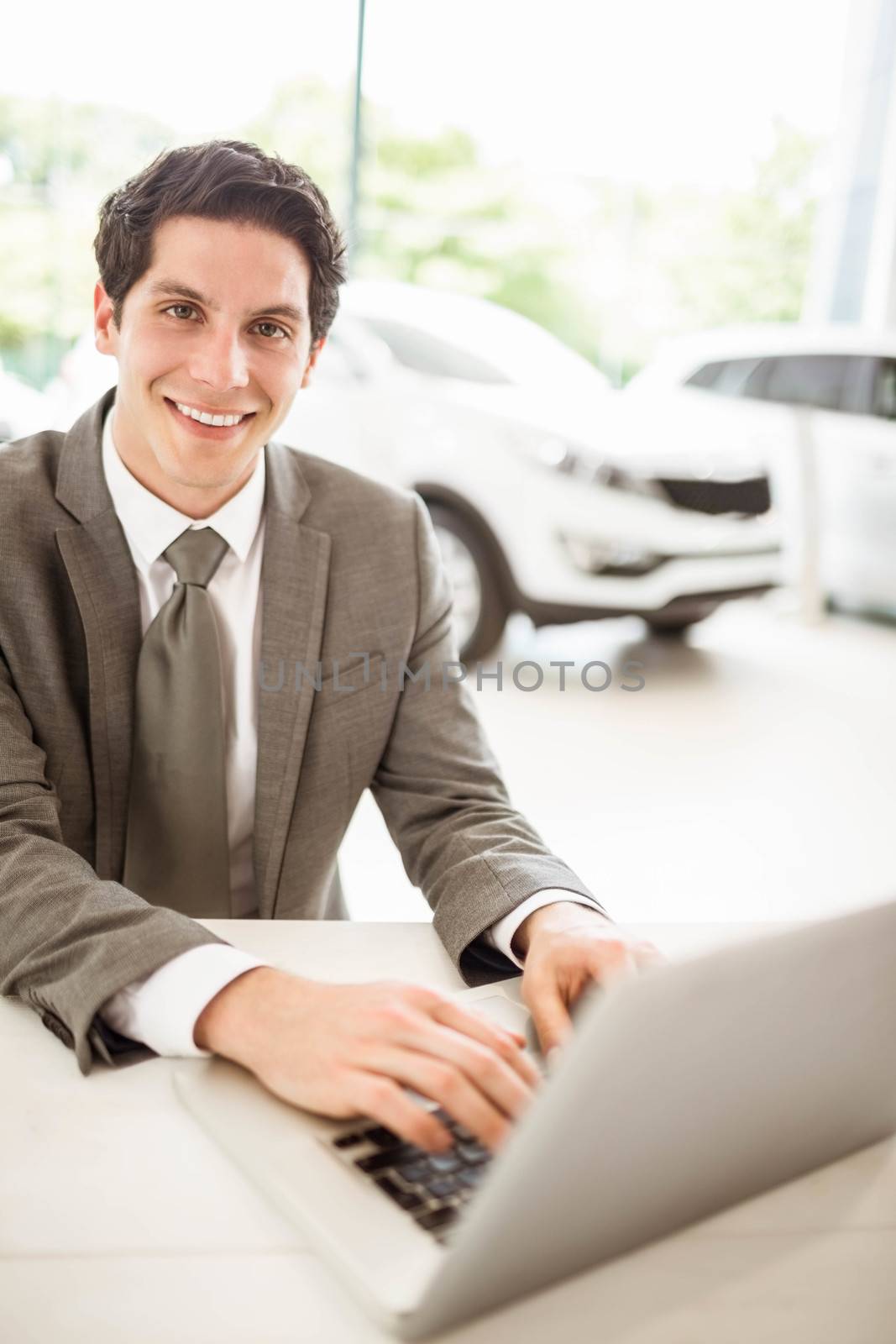 Smiling salesman typing on his laptop at new car showroom