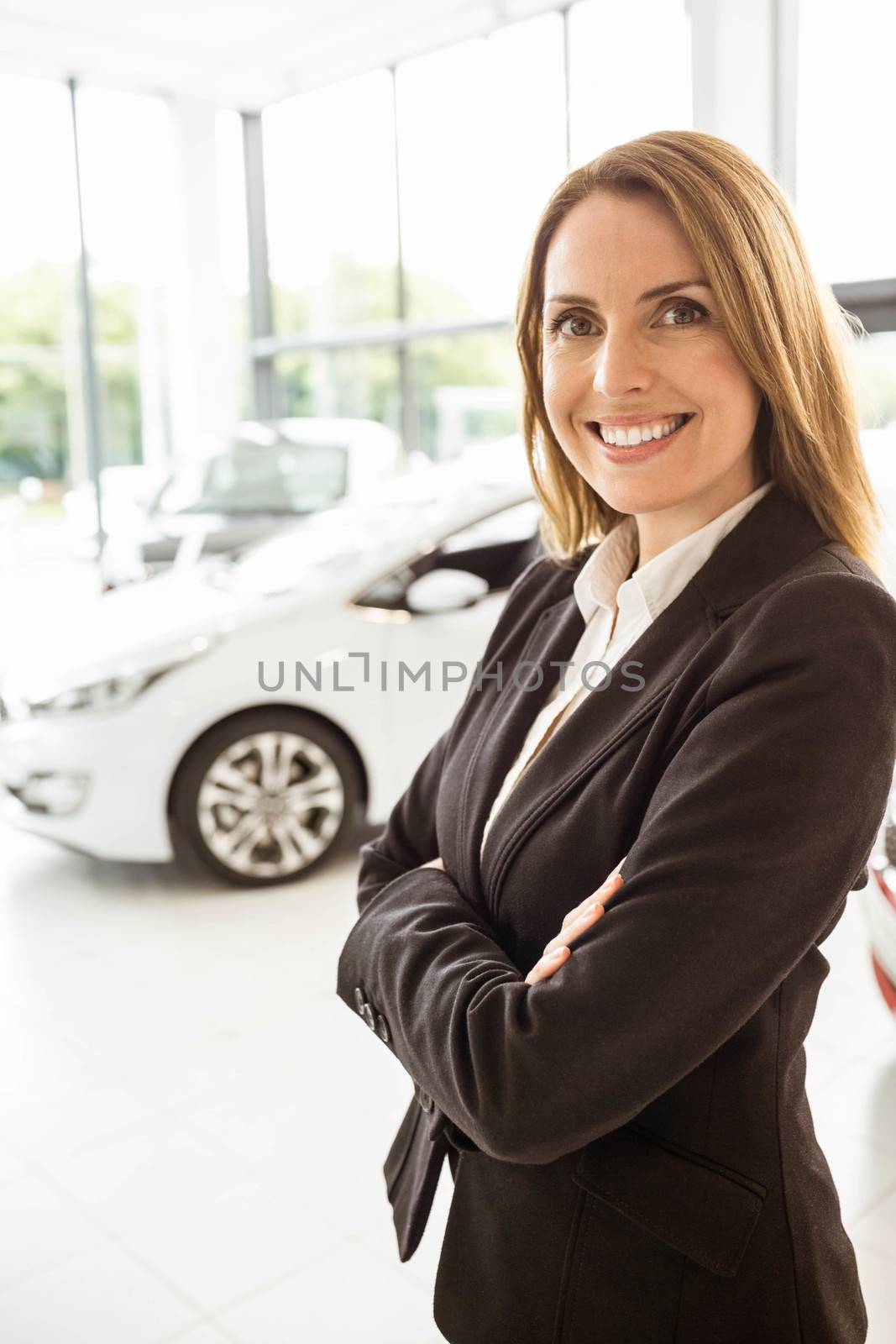 Smiling saleswoman standing with arms crossed at new car showroom