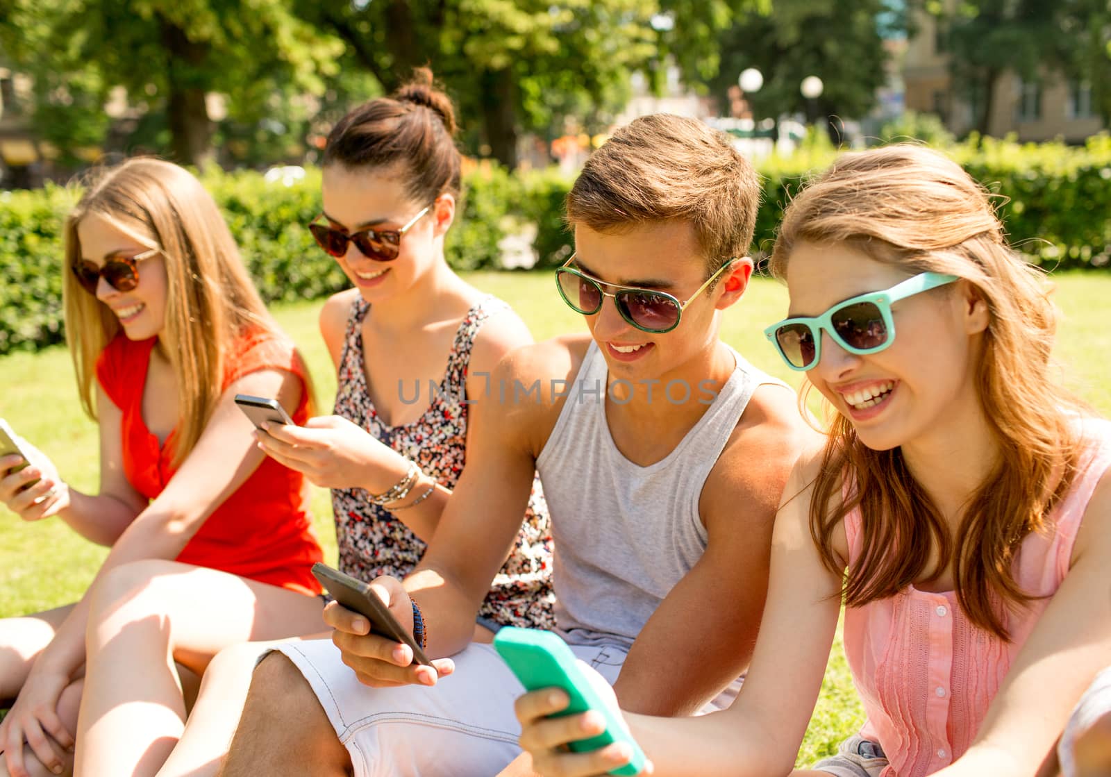 smiling friends with smartphones sitting on grass by dolgachov