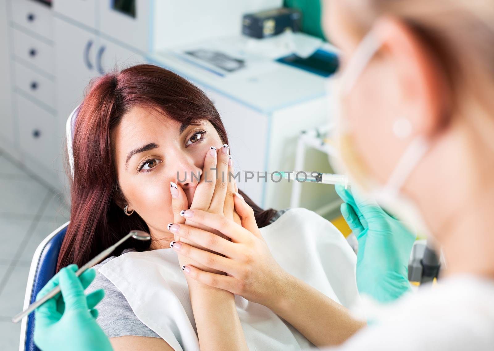 Young female dentist giving anesthesia to the patient before dental surgery. The patient in fear holds hands over mouth. Selective focus, focus on the patient.