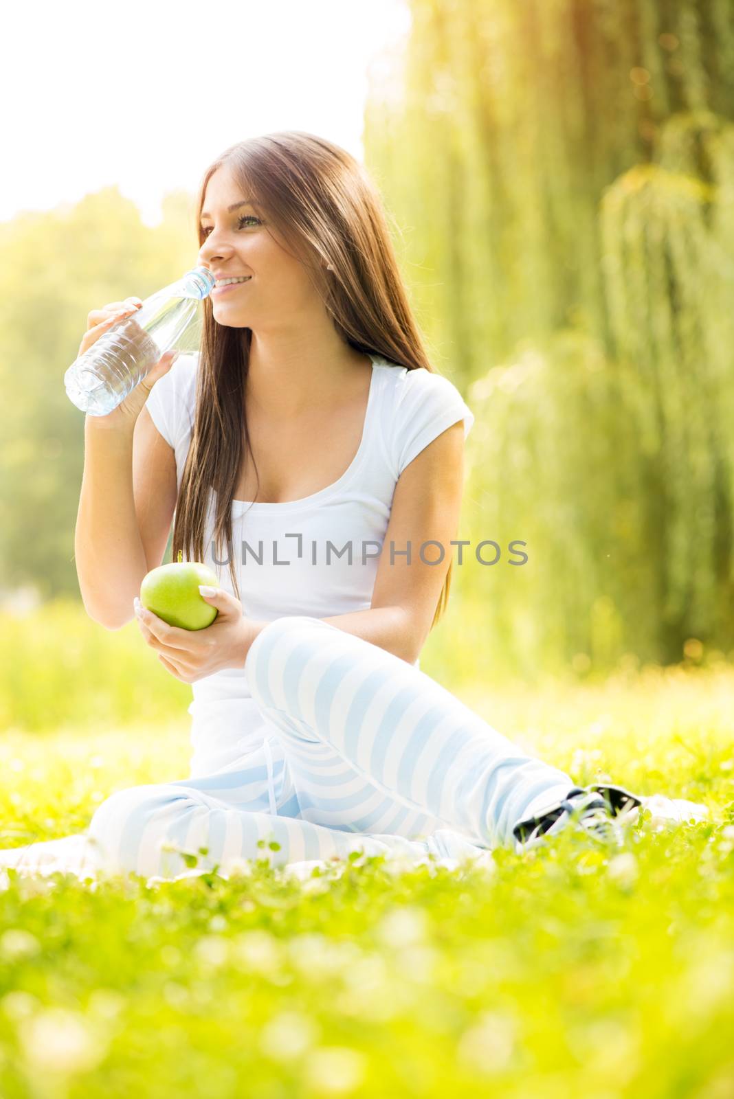 Smiling young woman holding an apple, drinks water from a bottle and enjoying in the nature.