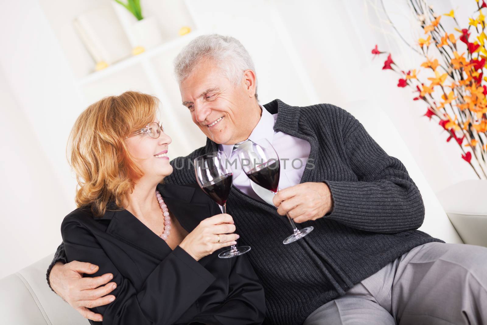 Happy senior couple sitting embraced at home, smiling and drinking red wine.