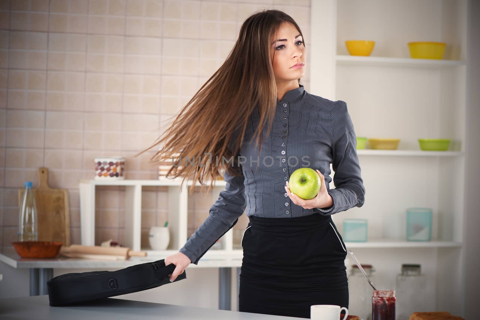 Businesswoman in the kitchen getting ready for work. She is rushing for work. Holding a bag with laptop in one hand and apple in other.