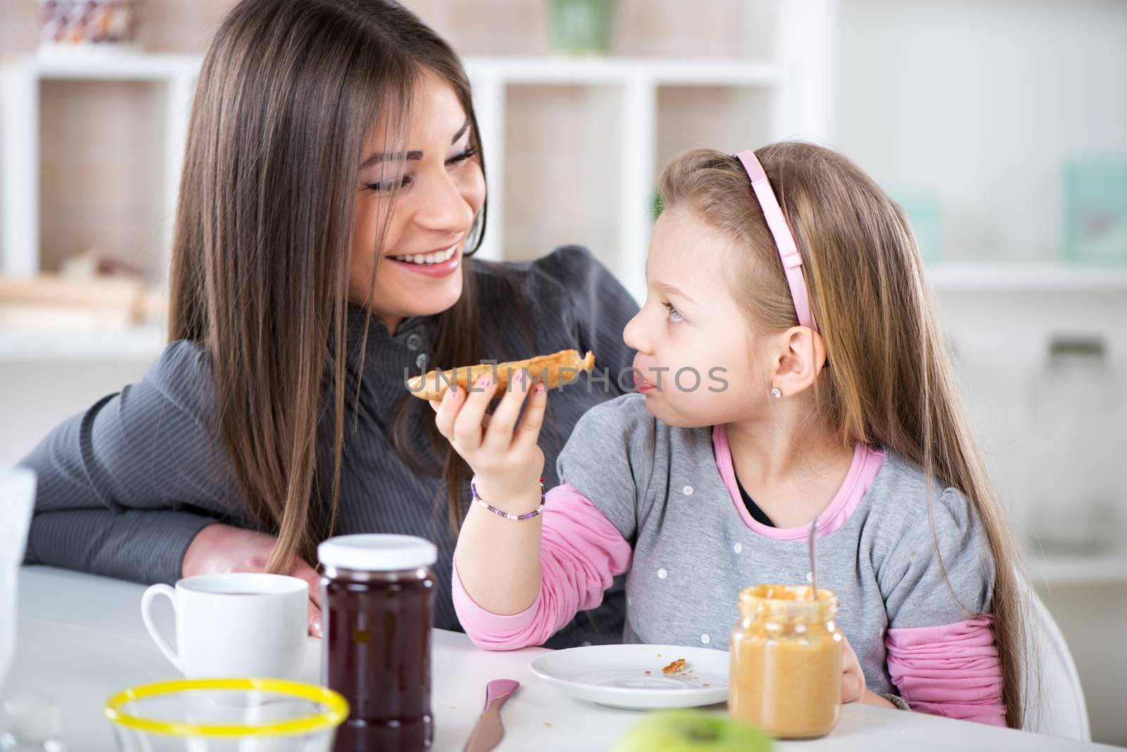 Mom and daughter breakfast in the kitchen environment and talking. Cute little girl eats bread with peanut butter.