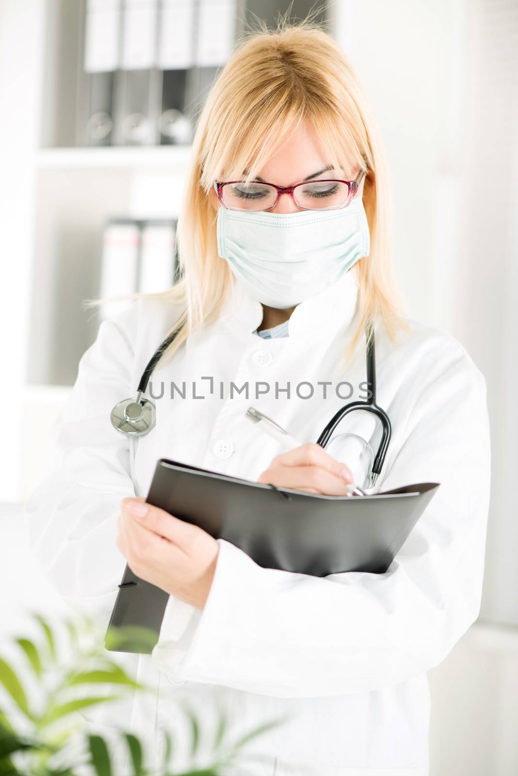 Portrait of a young woman doctor with stethoscope and surgical mask standing in her office and holding clipboard.