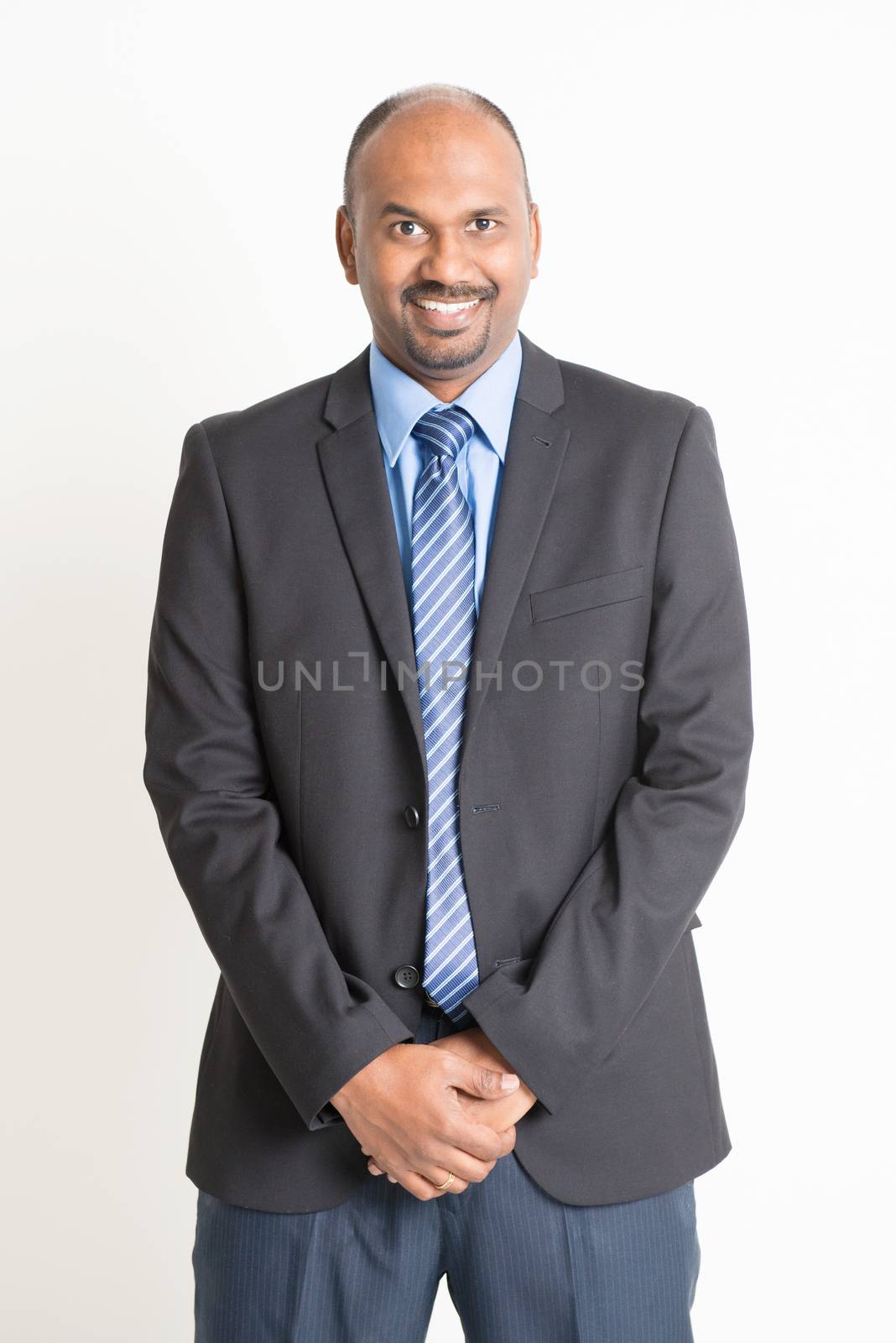 Friendly Indian businessman in formal suit looking at camera, standing on plain background.