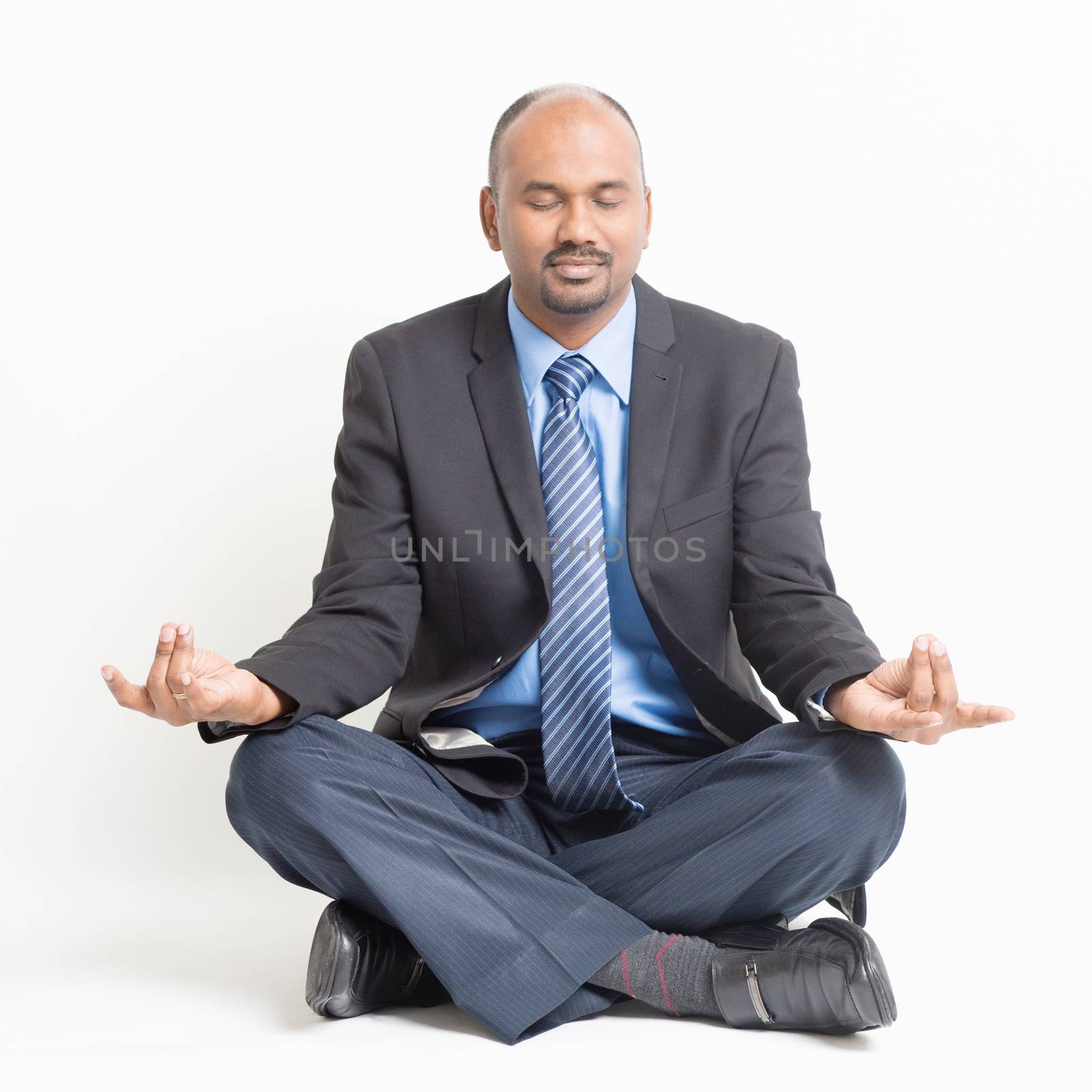 Business stress management concept. Full length Indian businessman in formal suit eyes closed sitting in meditation yoga pose, on plain background.