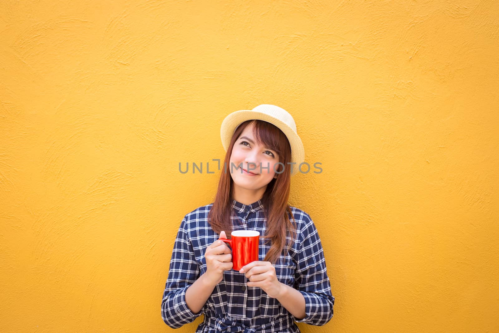 smiling woman wear in dress and hat holding red coffee cup on yellow cement wall background, thinking, imagination by powerbeephoto