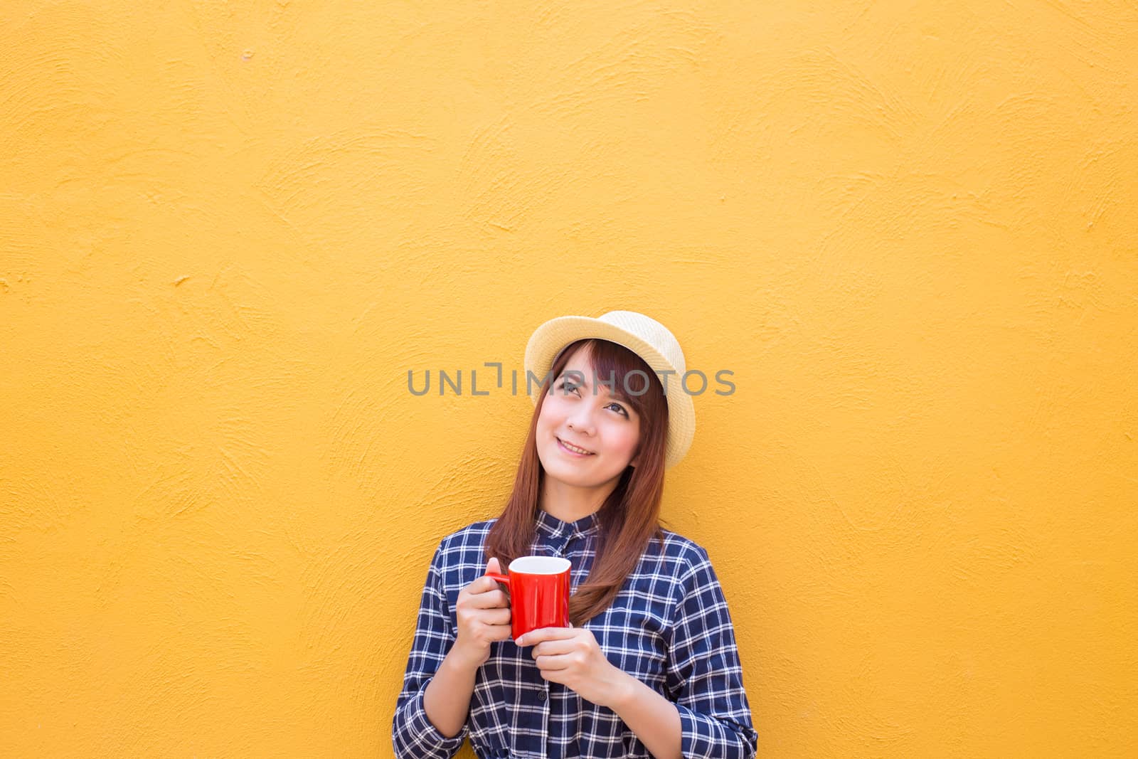 smiling woman wear in dress and hat holding red coffee cup on yellow cement wall background, thinking, imagination
