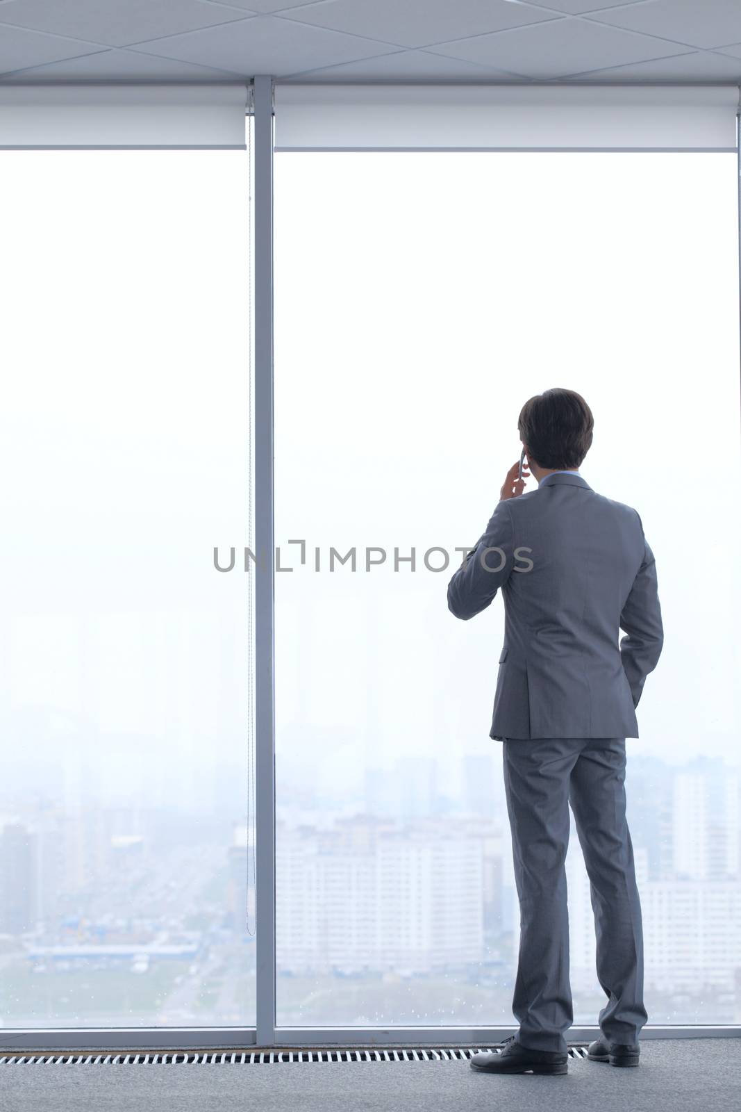 Businessman standing near window, looking at city and talking by phone