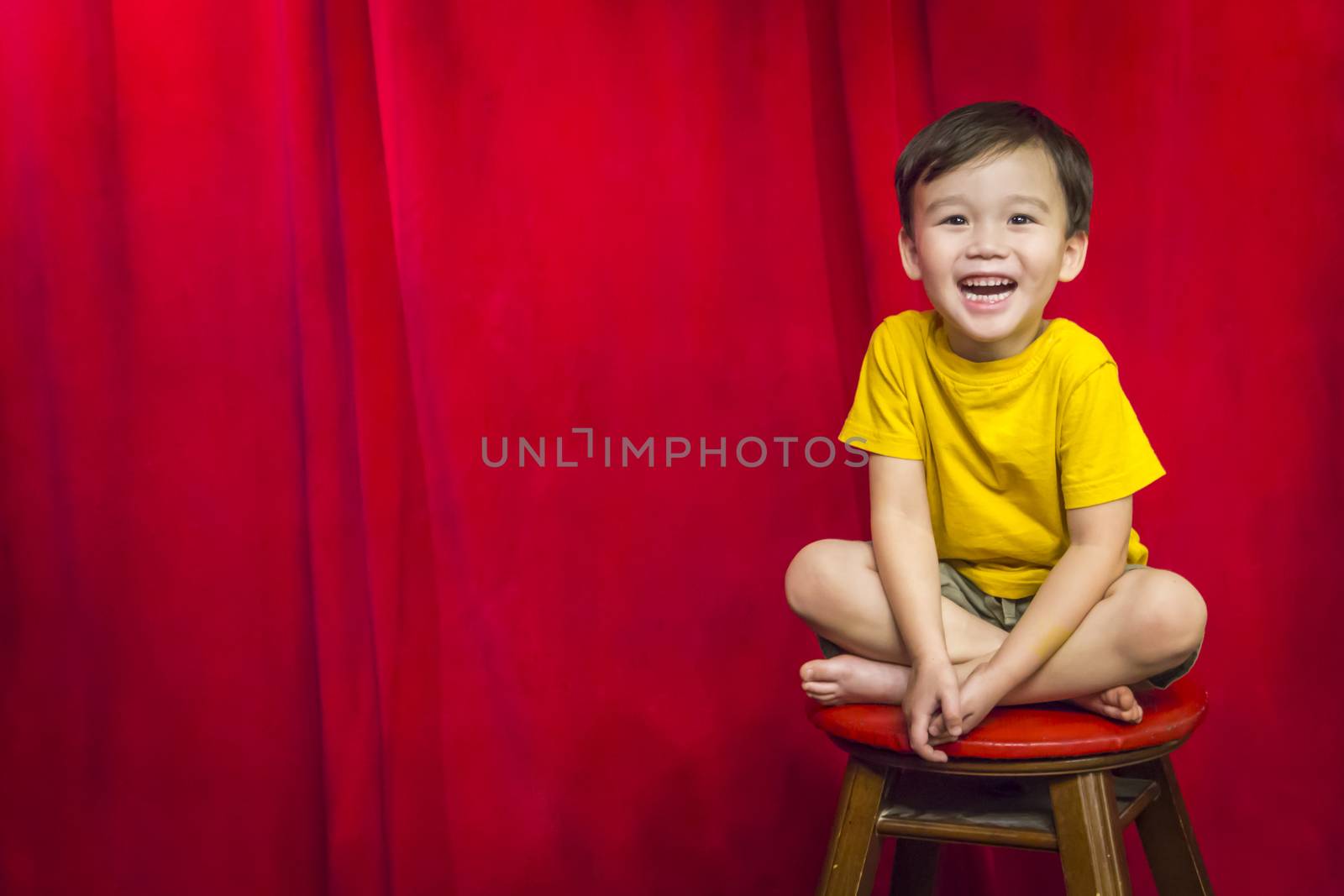 Laughing Mixed Race Boy Sitting on Stool in Front of Red Curtain.