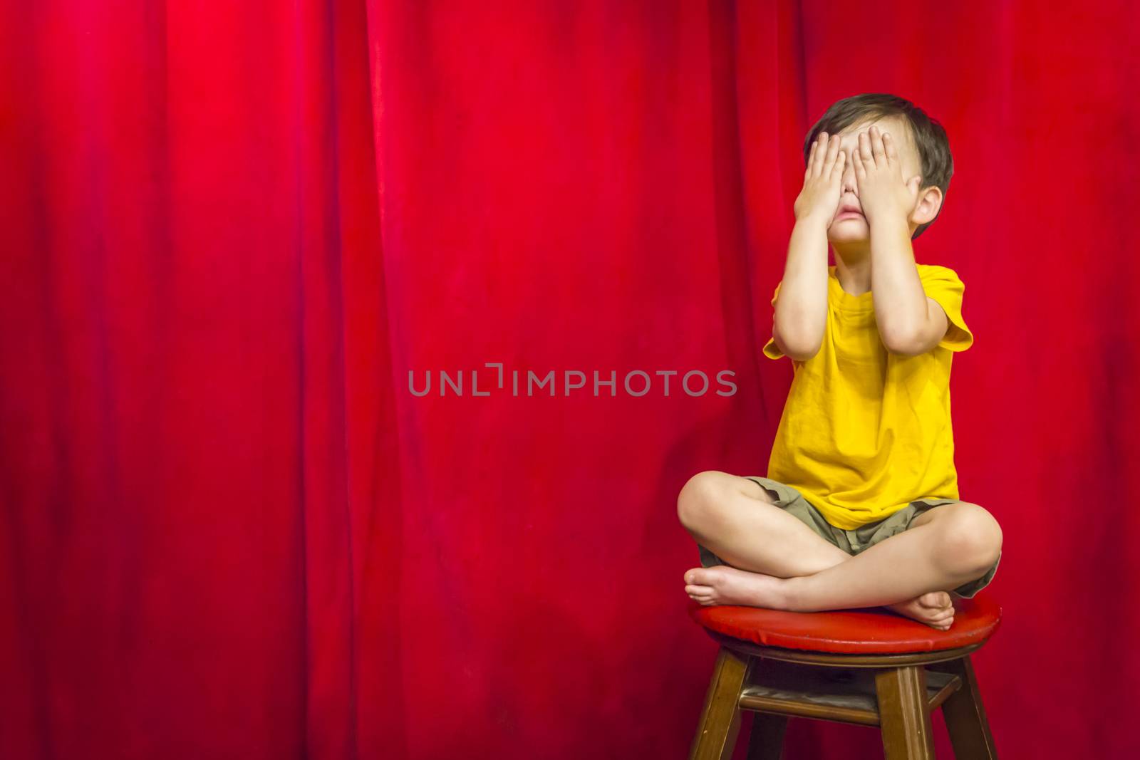 Boy Covering Eyes Sitting on Stool in Front of Curtain by Feverpitched