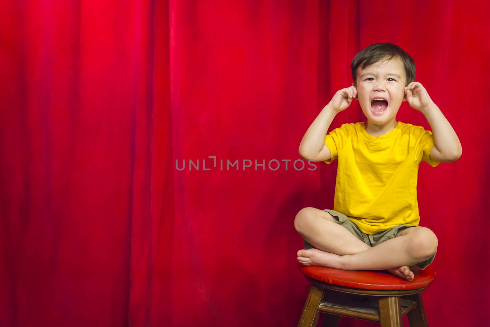 Mixed Race Boy With His Fingers In His Ears Sitting on Stool in Front of Red Curtain.