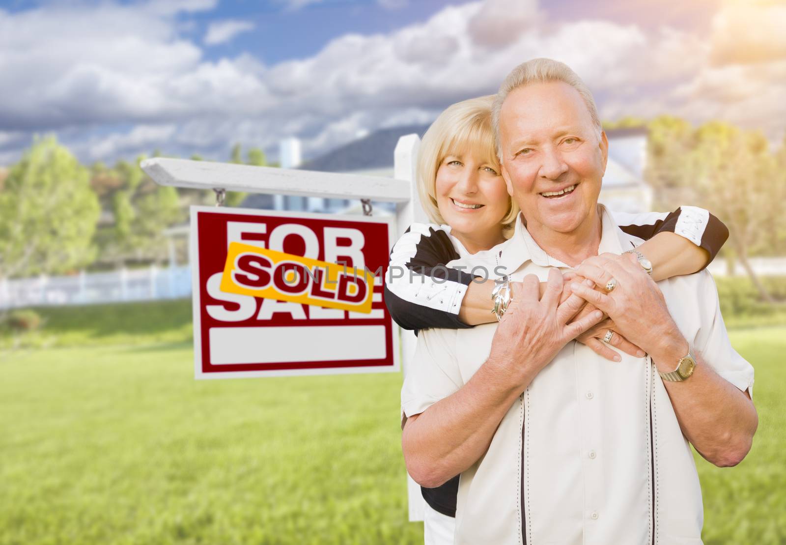 Happy Affectionate Senior Couple Hugging in Front of Sold Real Estate Sign and House.