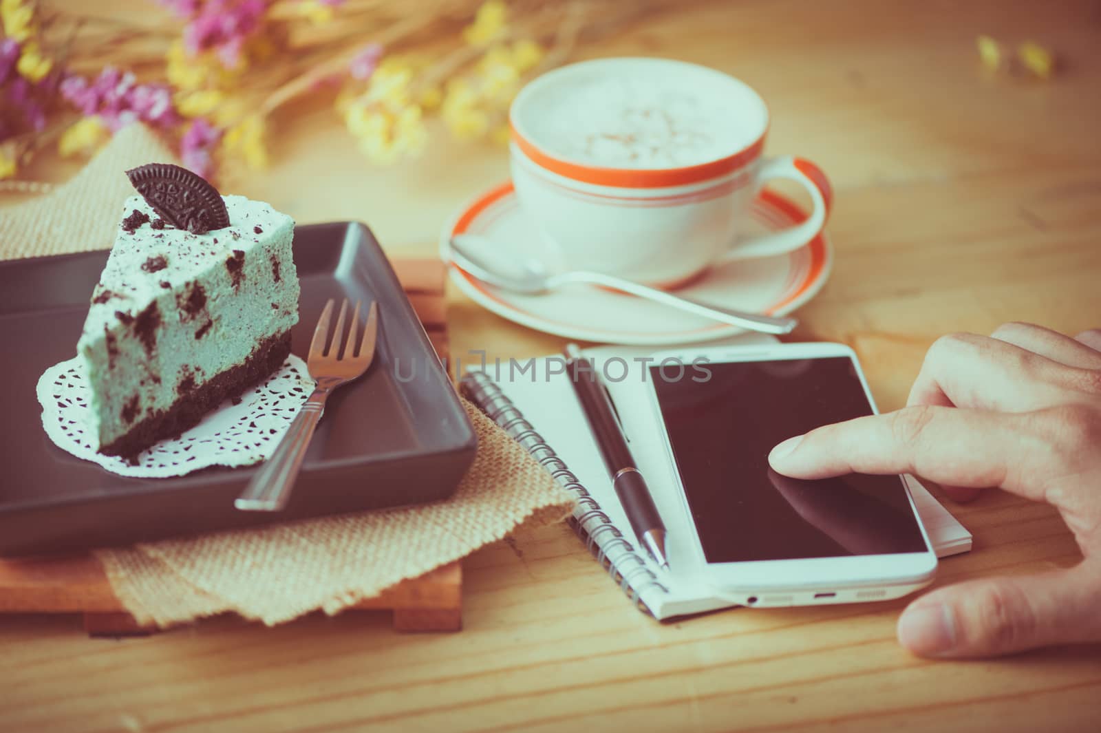 Male right hand finger touching on smartphone, peppermint with chocolate cookie cheese cake, notebook and pen, and hot cappuccino at cafe in afternoon time with film filter effect