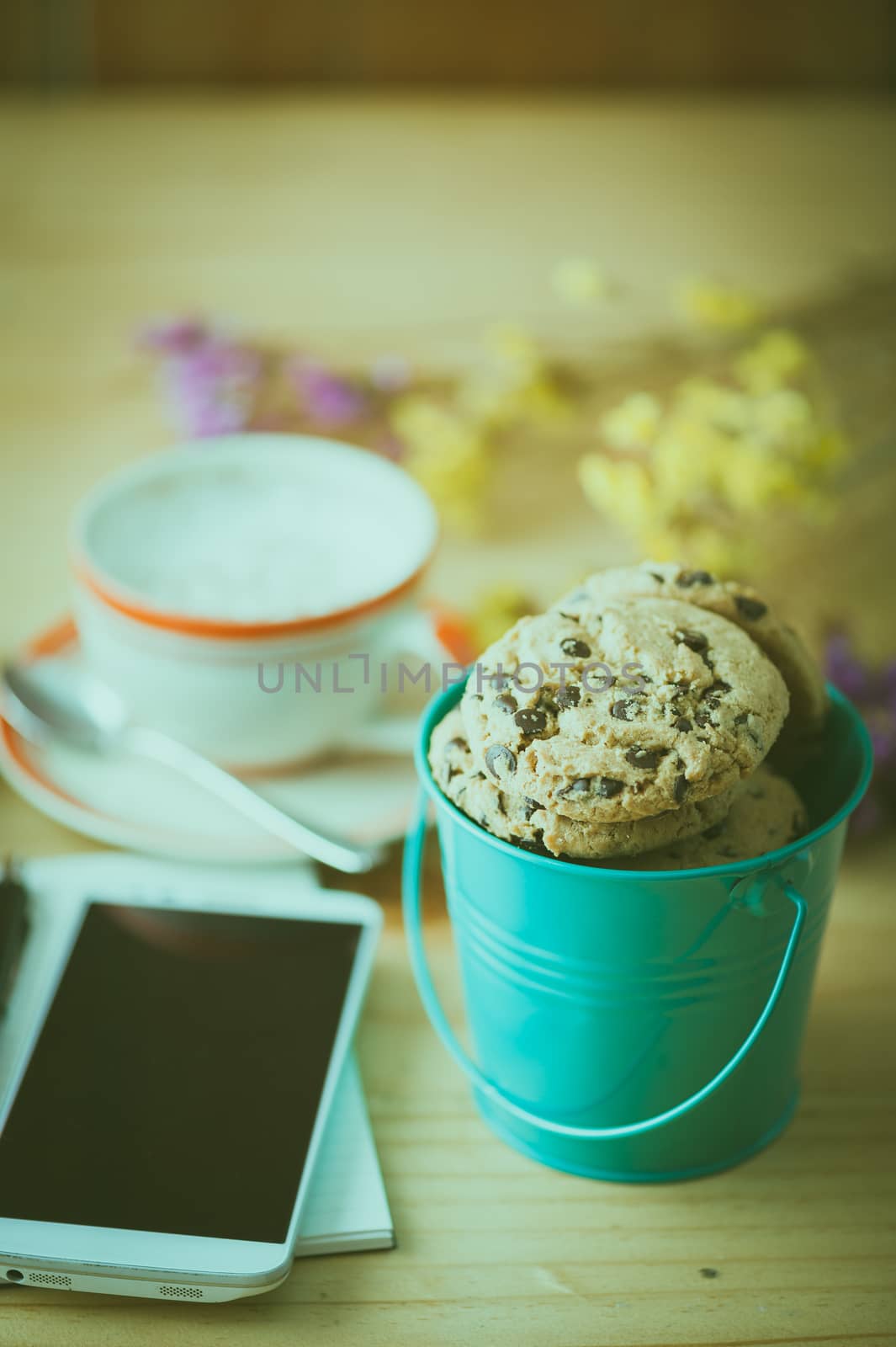 Chocolate chip cookies in small green bucket with smartphone, notebook and pen, and coffee cup at cafe in morning time with film filter effect