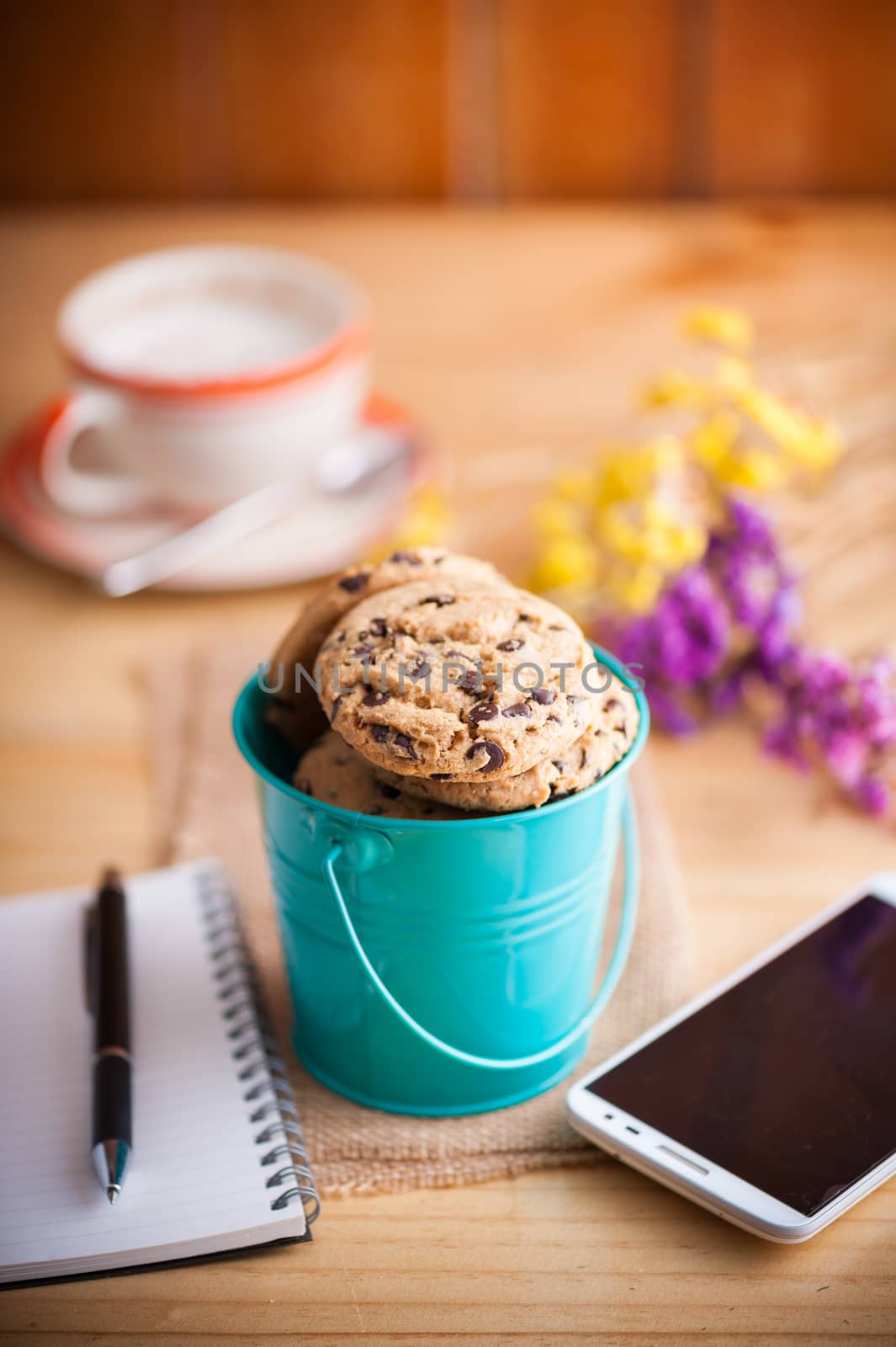 Chocolate chip cookies in small green bucket with smartphone, notebook and pen, and coffee cup at cafe in morning time