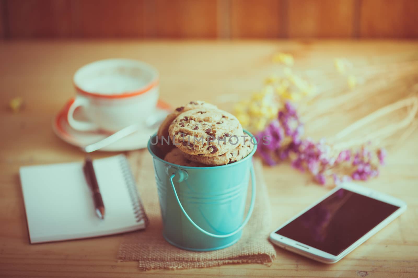 Chocolate chip cookies in small green bucket with smartphone, notebook and pen, and coffee cup at cafe in morning time with film filter effect