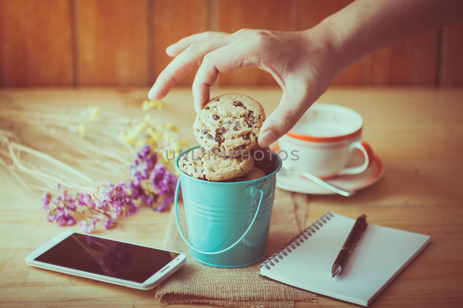Male right hand taking chocolate chip cookie from small green bucket with smartphone, notebook and pen, and coffee cup at cafe in morning time with vintage filter effect