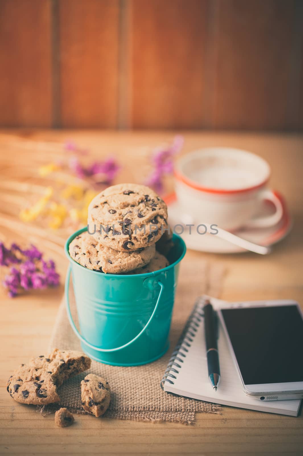 Chocolate chip cookies in small green bucket with smartphone, notebook and pen, and coffee cup at cafe in morning time with vintage filter effect