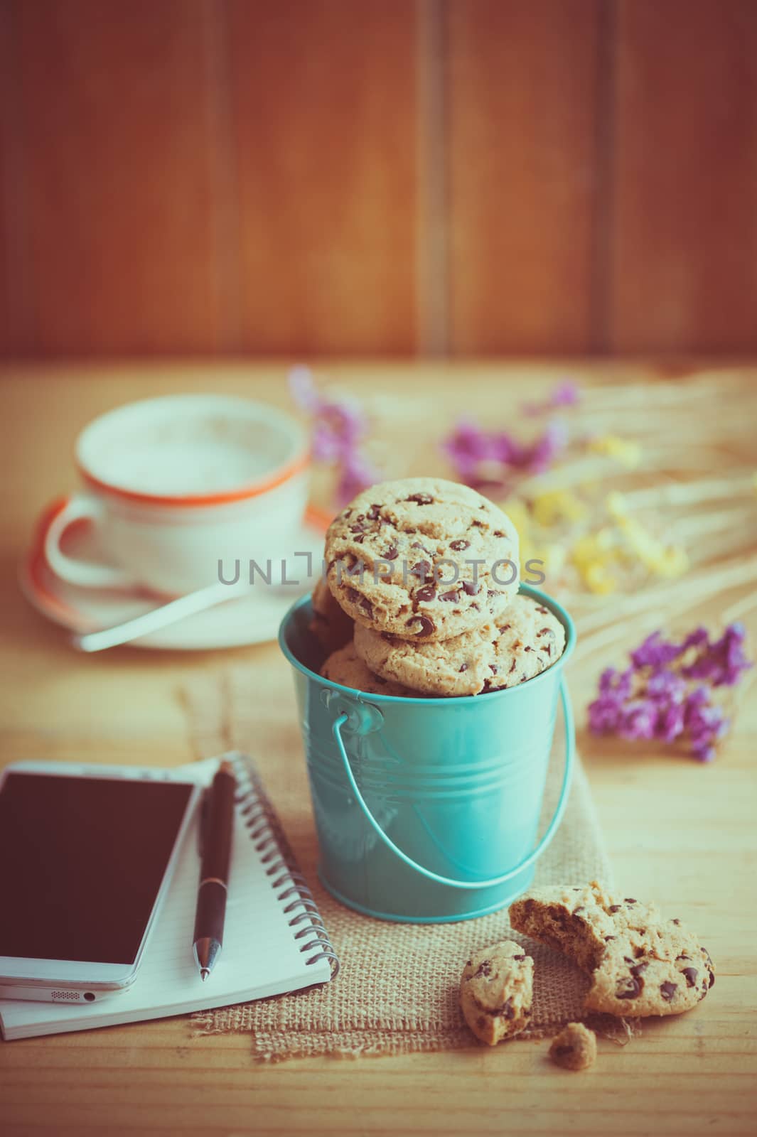 Chocolate chip cookies in small green bucket with smartphone, notebook and pen, and coffee cup at cafe in morning time with vintage filter effect