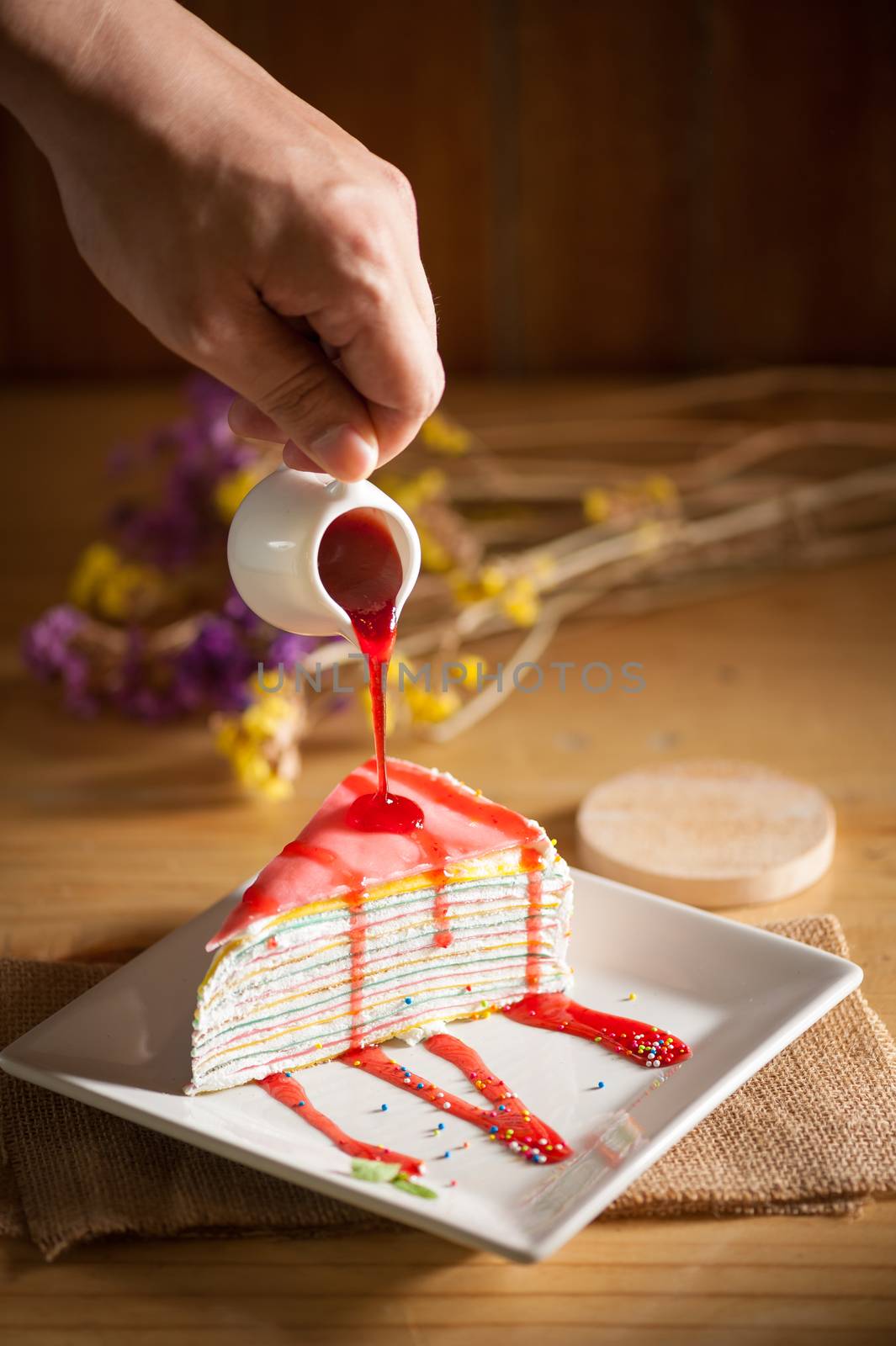 Rainbow crepe cake with male left hand pouring strawberry sauce on top in afternoon time with low key scene