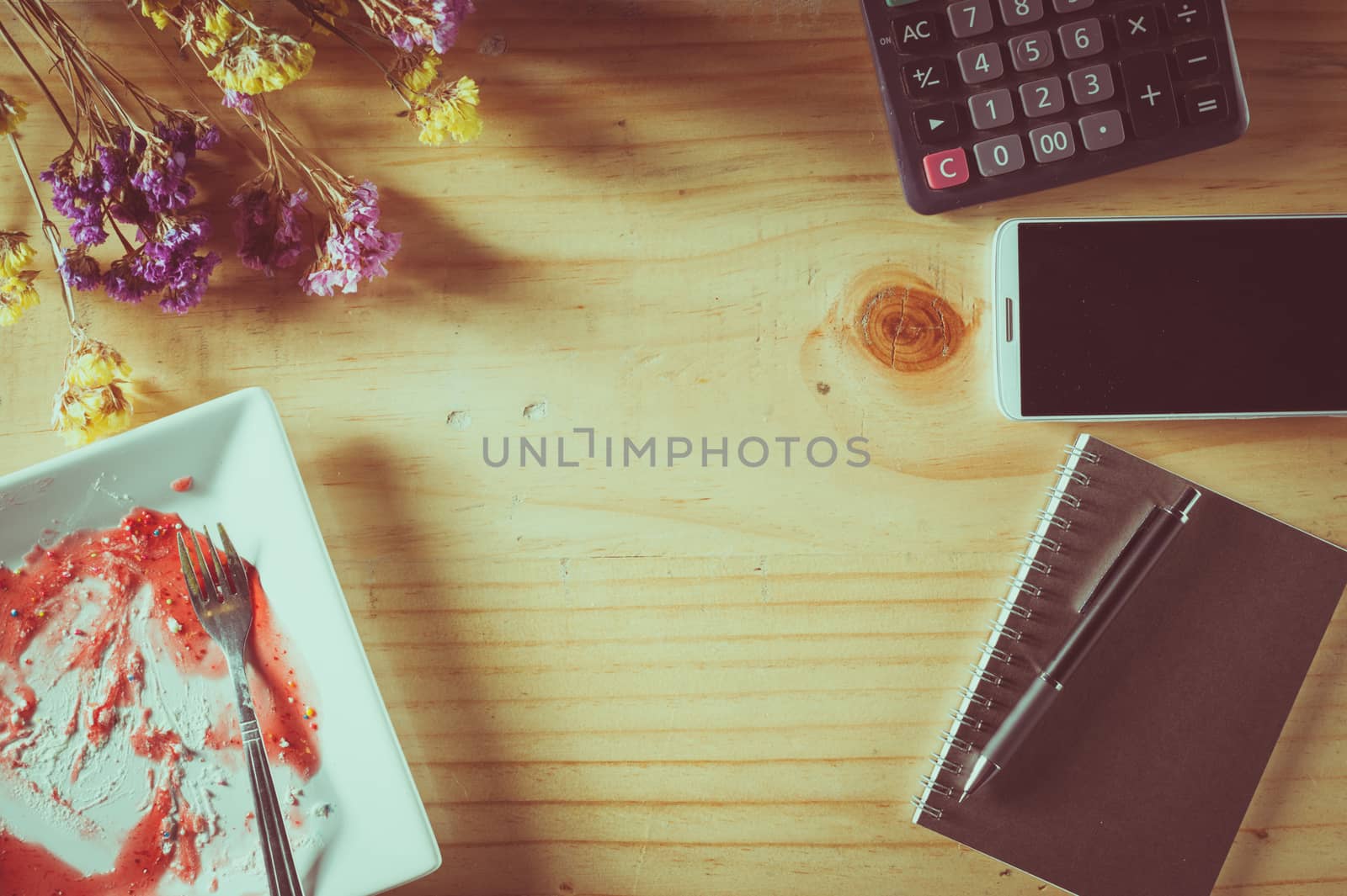 Wood table background with blank area for text or message, dirty dessert plate, flowers, calculator and smartphone as frame around in afternoon time with film filter effect