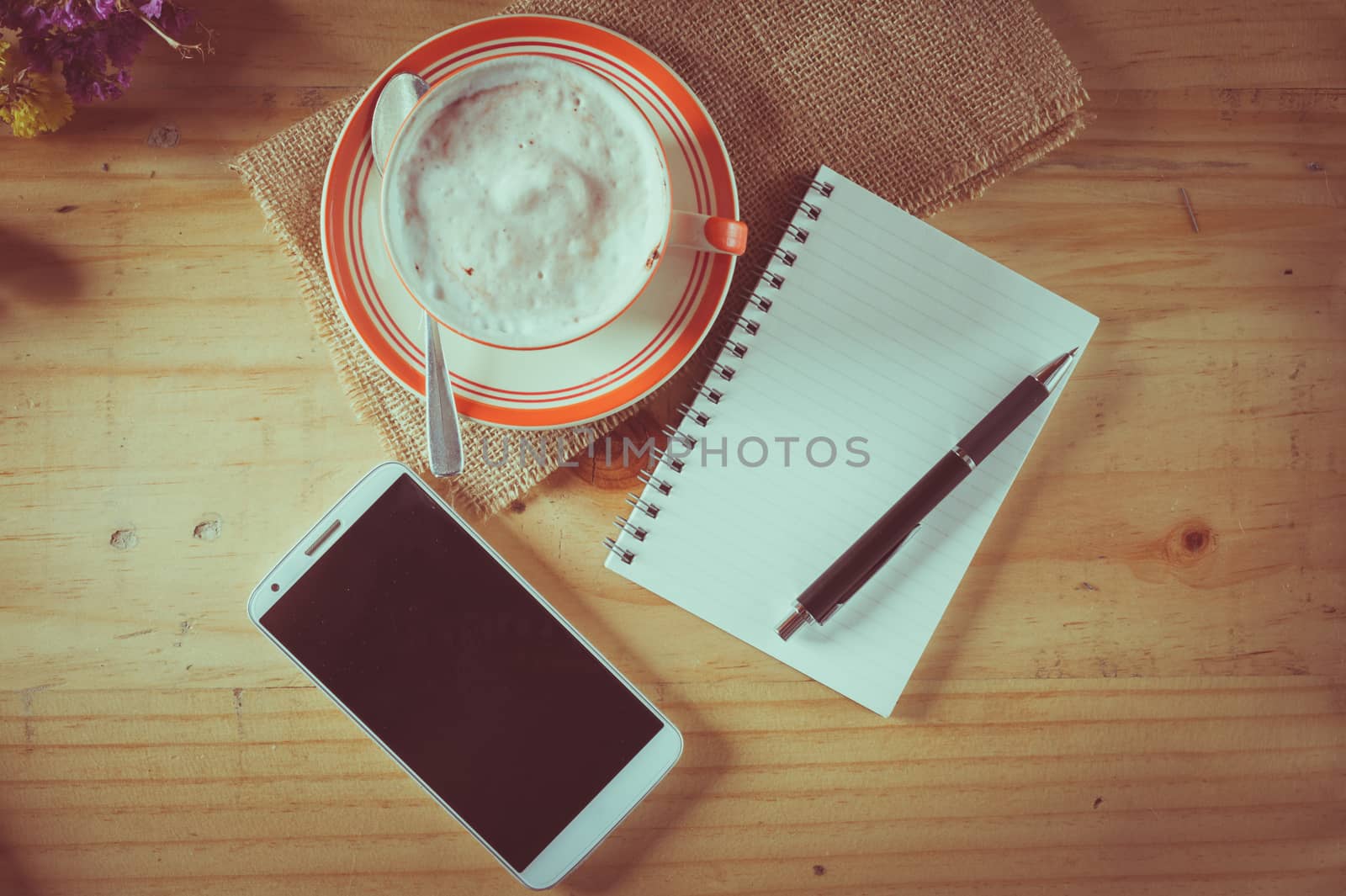 Opened notepad with blank area for text or message, pen, smart phone, and a cup of hot coffee on wood table in afternoon time with film filter effect