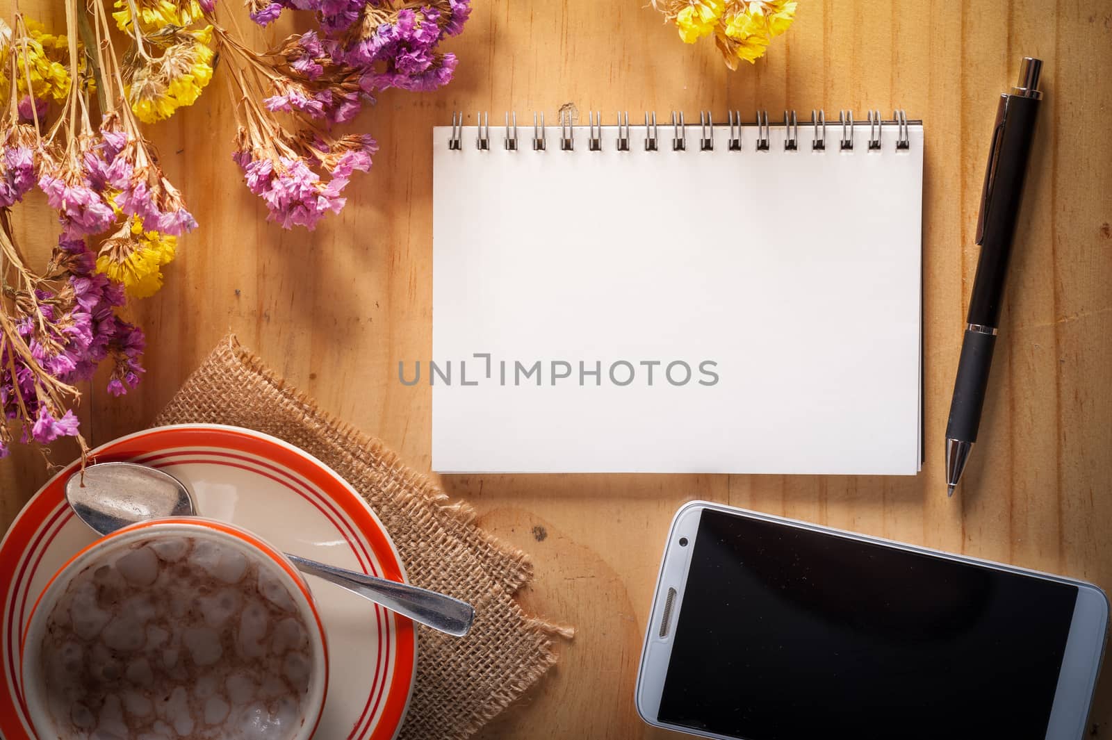 Opened notepad with blank area for text or message, pen, smart phone, and a cup of hot coffee on wood table in afternoon time