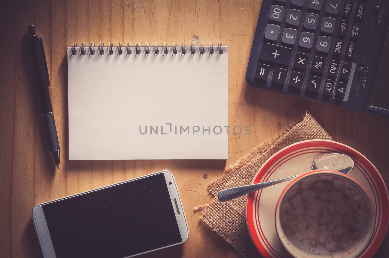 Opened notepad with blank area for text or message, pen, smart phone, and calculator on wood table in evening time with film filter effect