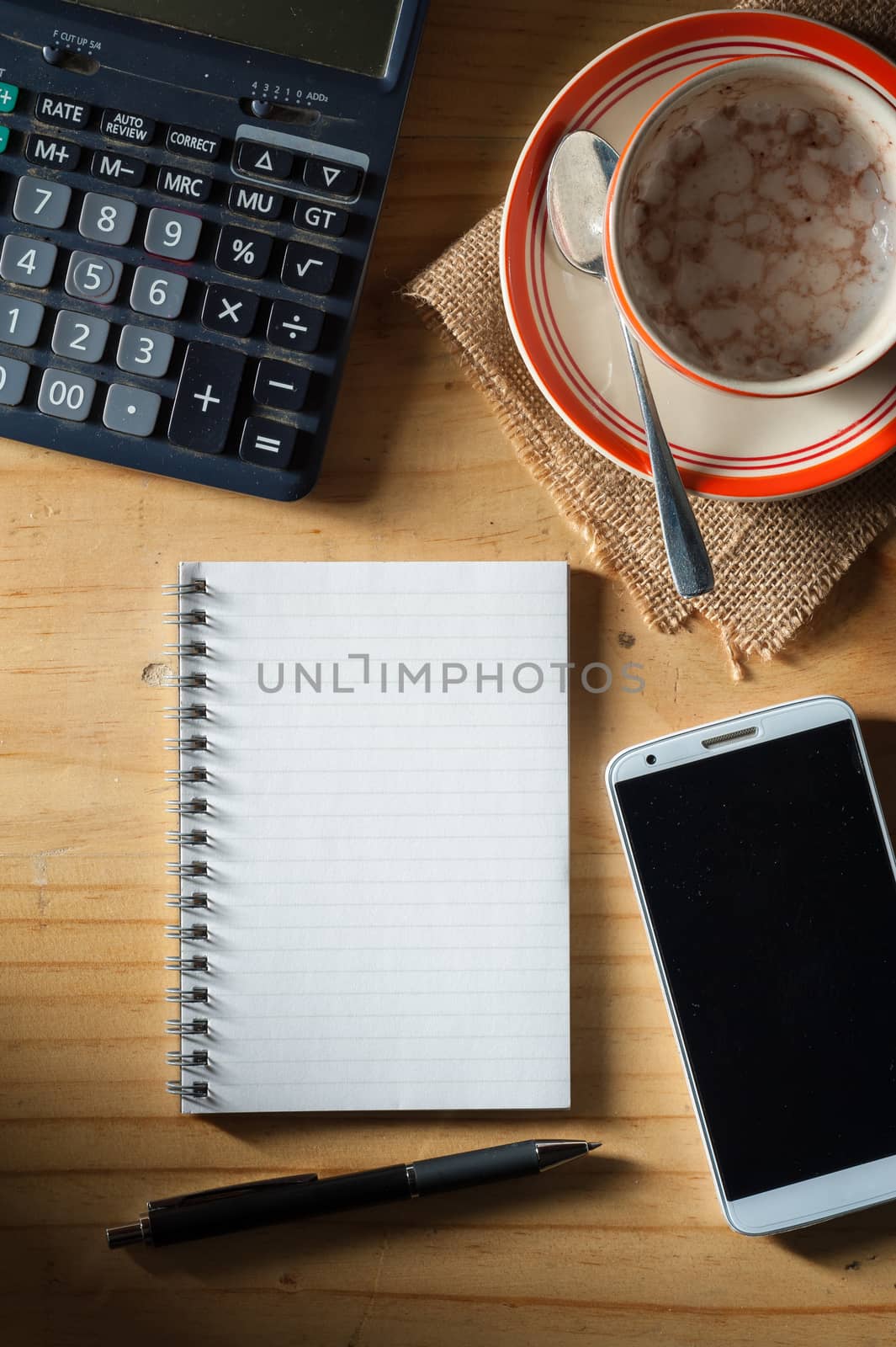 Opened notepad with blank area for text or message, pen, smart phone, and calculator on wood table in evening time with low key scene