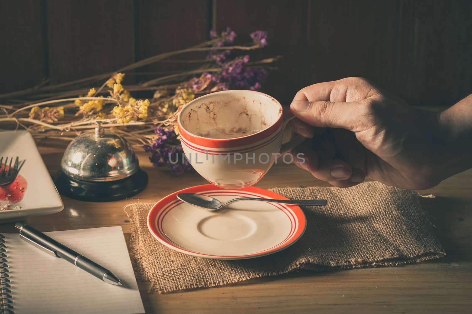 Male right hand holding a cup of coffee in cafe with notebook and pen on table in morning time with low key scene and film filter effect