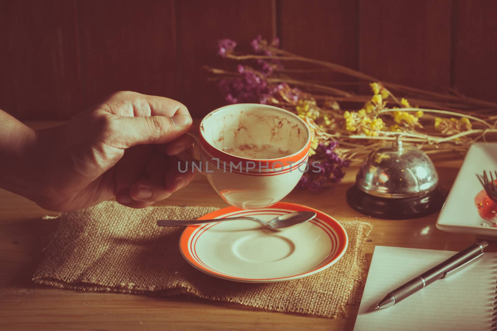 Male left hand holding a cup of coffee in cafe with notebook and pen on table in morning time with low key scene and film filter effect
