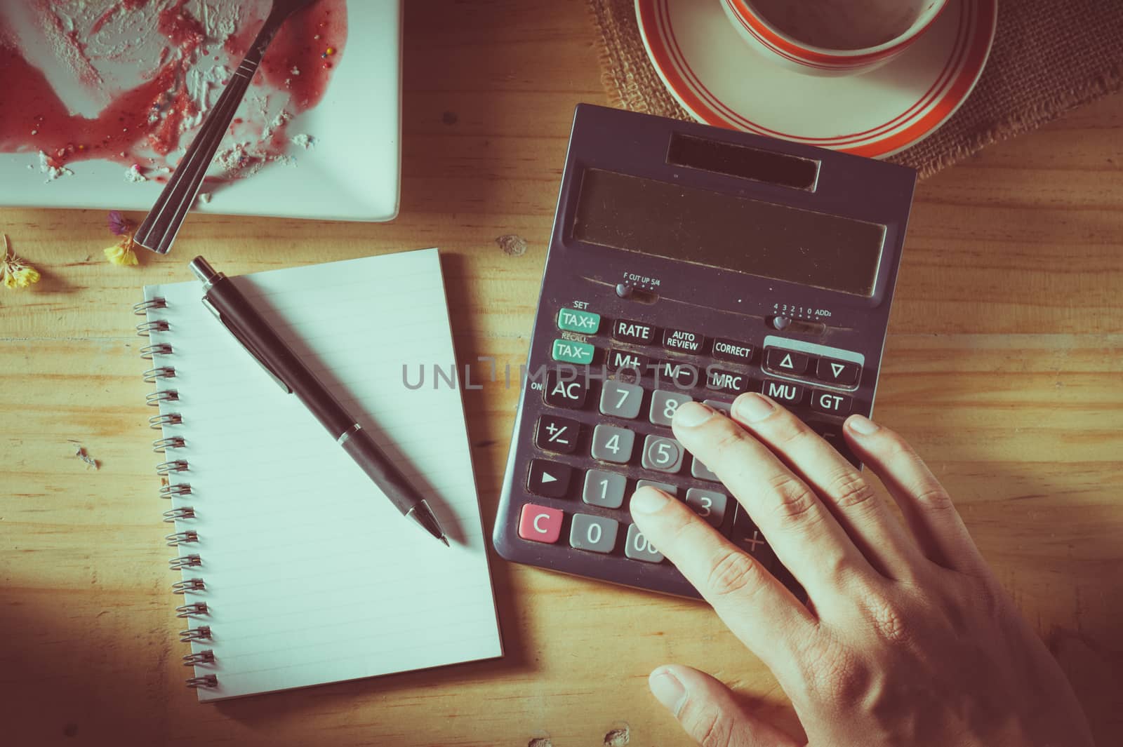 Male hand using calculator with opened notepad, pen, smart phone, and calculator on wood table in evening time with low key scene and film filter effect