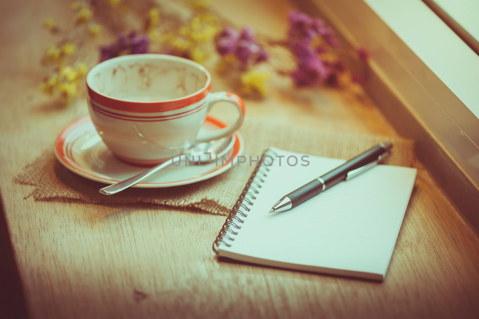 Coffee cup, opened notebook, and pen on wood bar beside window at coffee shop in afternoon time with film filter effect