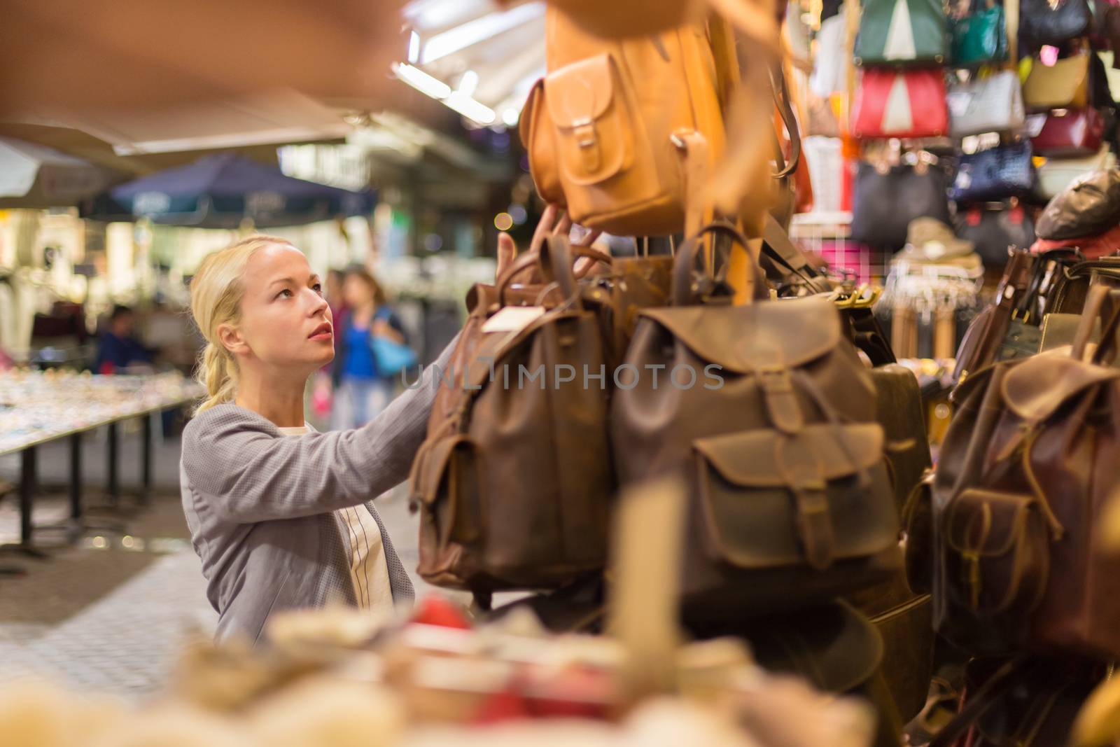 Beautiful casually dressed caucasian blond woman shopping for new leather bag on crafts market in Chania, Crete, Greece.