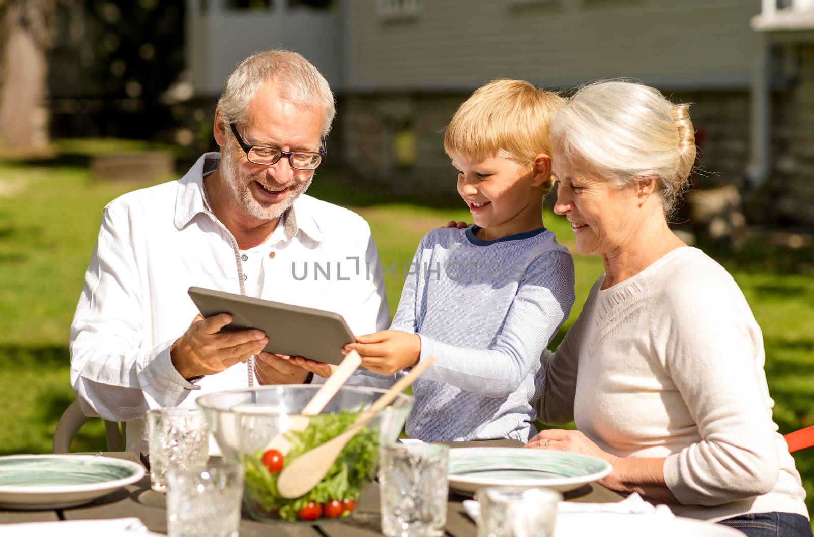 family, happiness, generation, home and people concept - happy family sitting at table with tablet pc computer outdoors