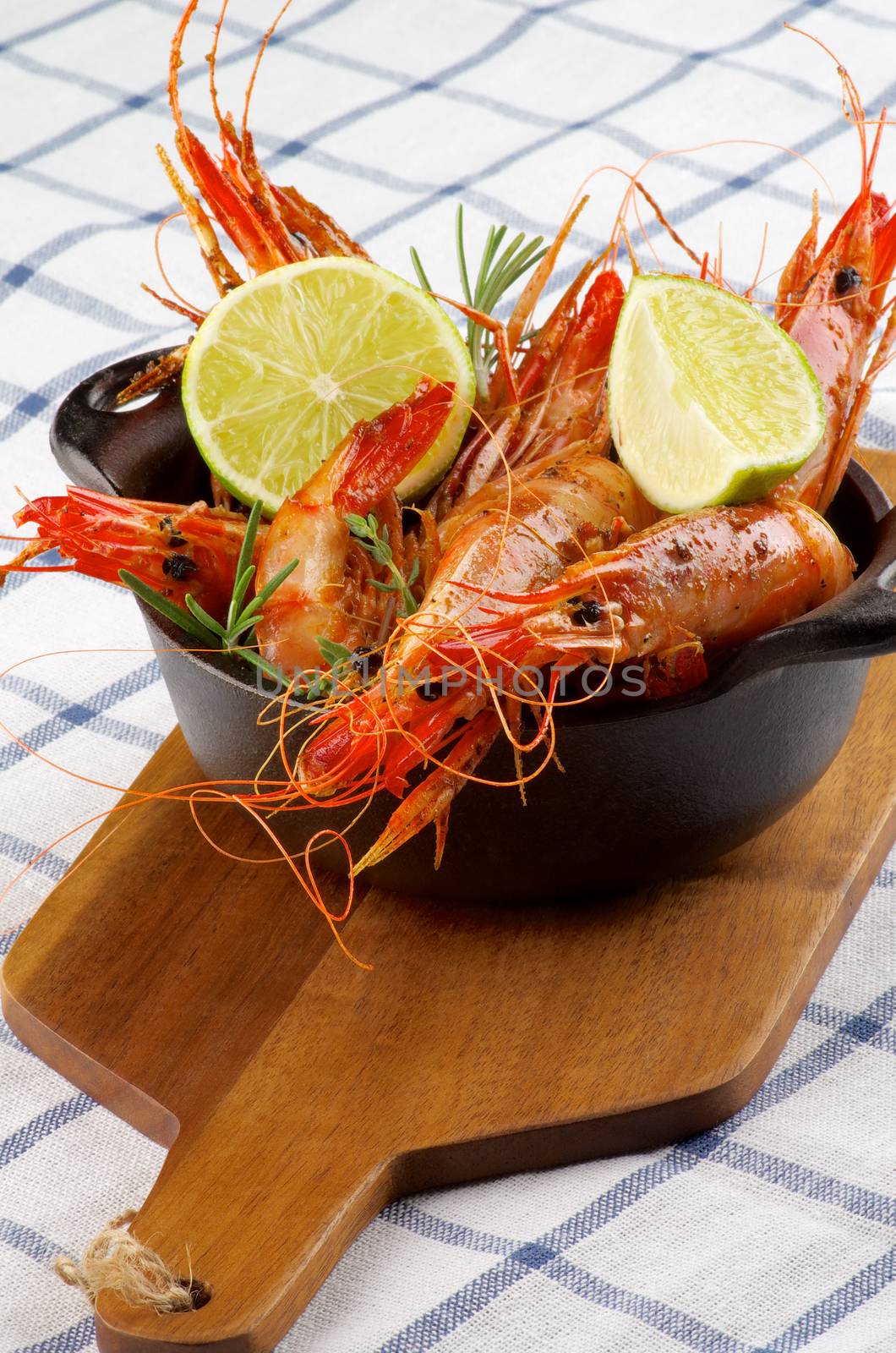 Delicious Roasted Shrimps in Cast Iron with Lime and Rosemary on Wooden Cutting Board closeup on Checkered Napkin background