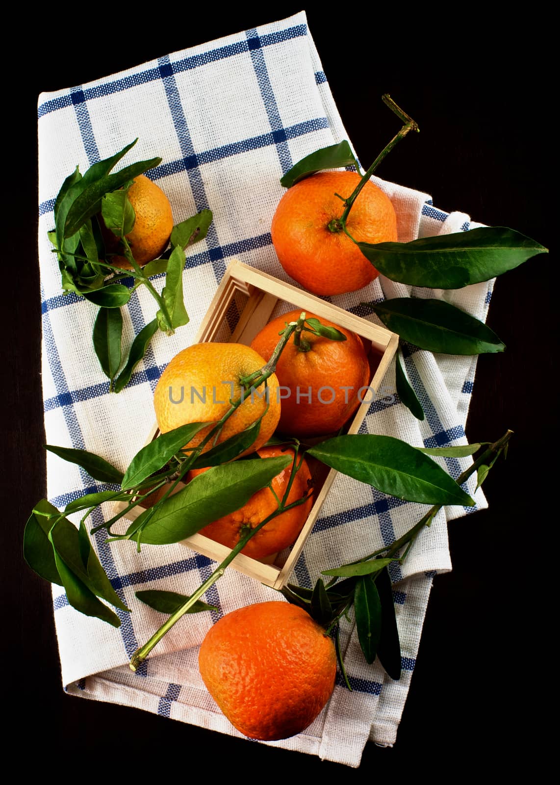 Ripe Tangerines with Leafs in Wooden Box on Napkin closeup on Dark Wooden background. Top View