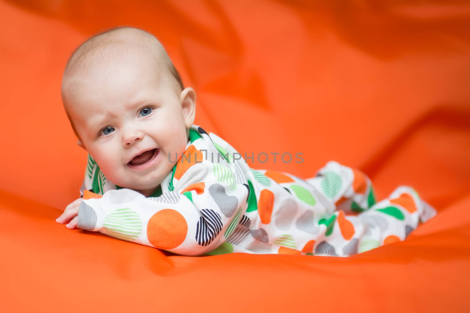 Baby girl in a white pjama with big dots lying on a belly on an vibrant orange pillow