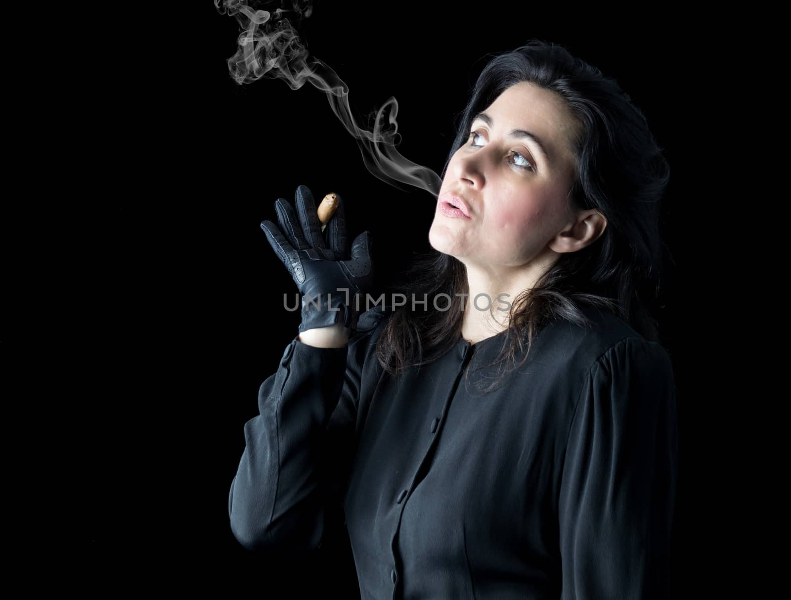 Brunette woman in black dress and black gloves standing in front of a black backdrop, holding a cigar in one hand and blowing smoke.