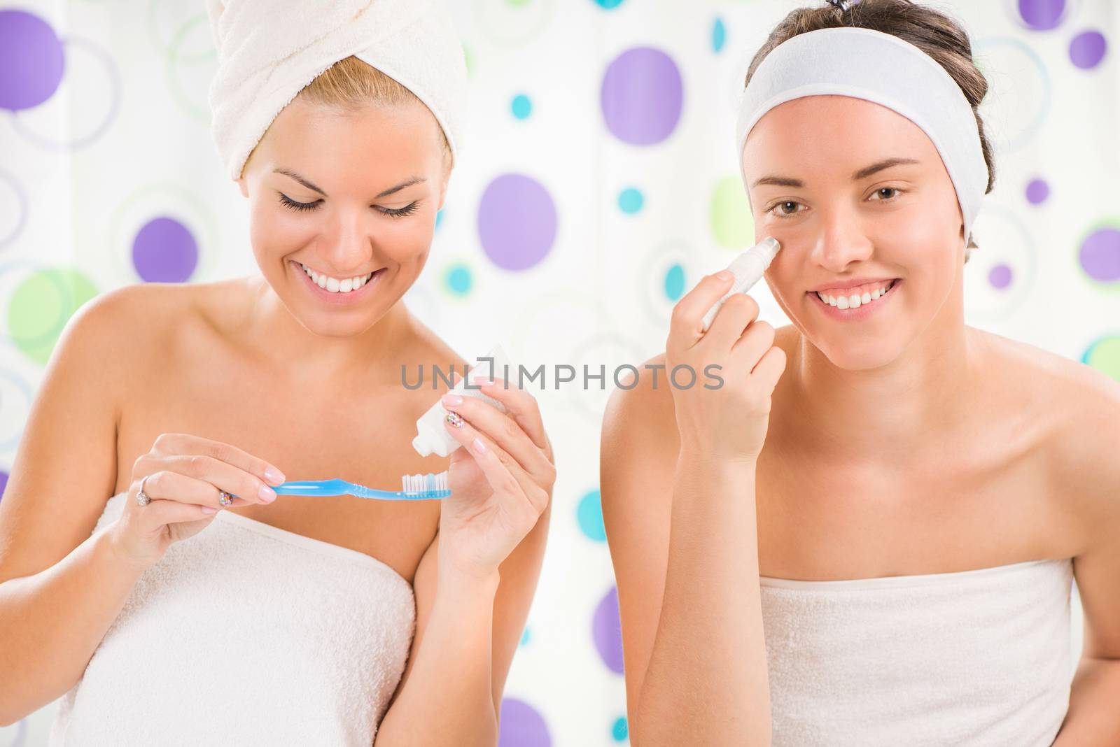 Two young cute woman preparing to start their day. One girl brushing teeth, the other girl applying cream.